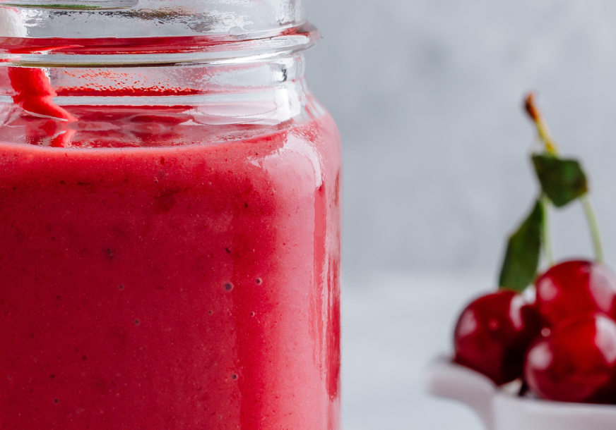 Glass jar filled with a red smoothie with a trio of cherries in the background